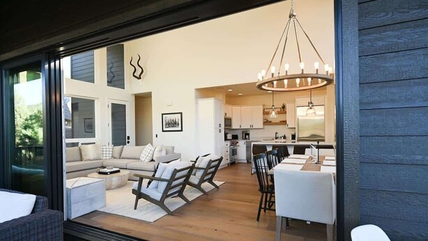 A modern living room with a sofa, chairs, and a dining table. There is a kitchen in the background with a chandelier.