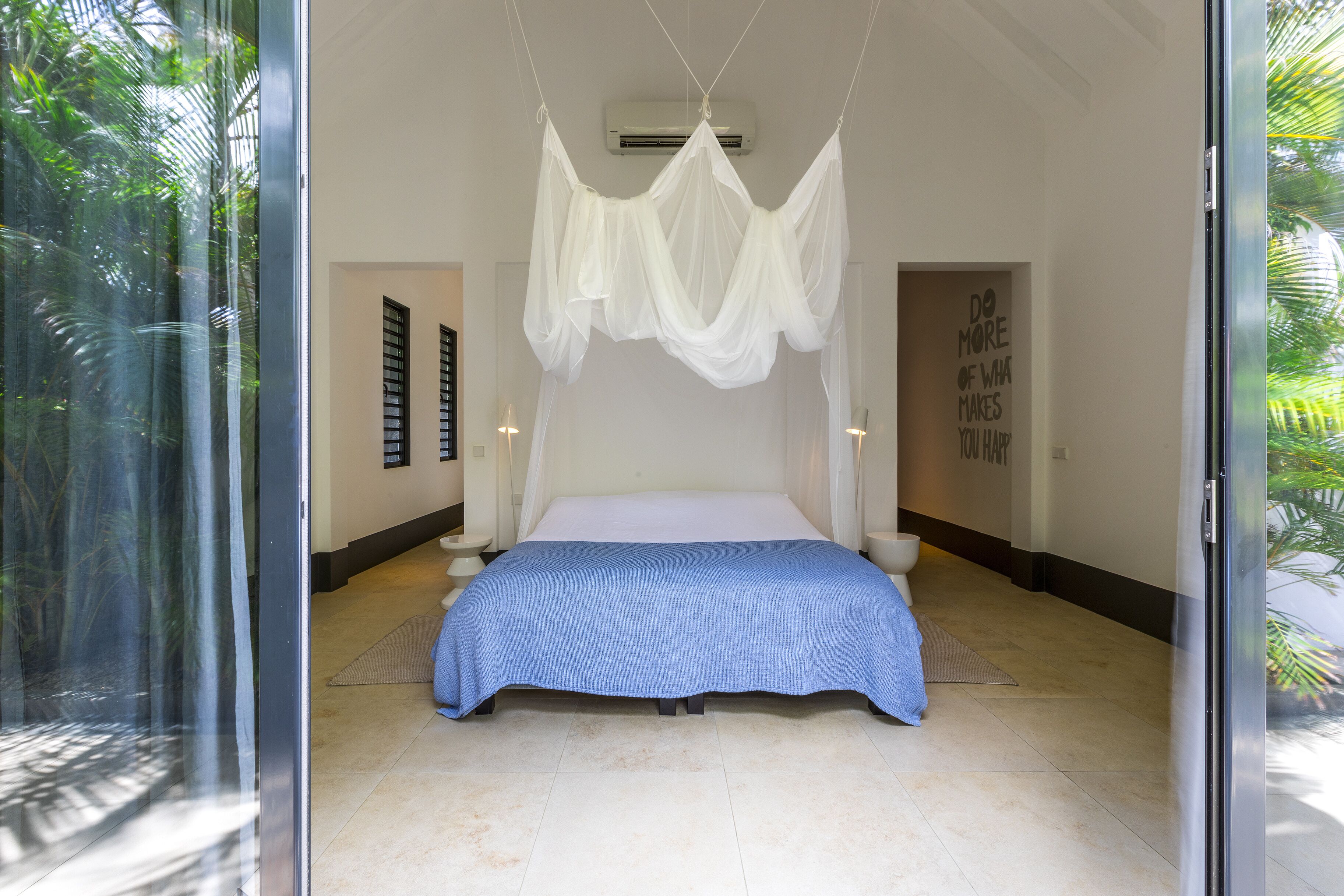 Un dormitorio con cama, dosel, pared con una cita motivacional y vistas a la vegetación a través de un ventanal.