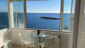 A room with a glass table and two chairs, a coffee maker, and a view of the sea and a small island.