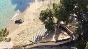 A beach with clear water, a sandy shore, and a few trees. There are buildings in the background.
