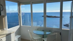 A room with a glass table and two chairs, a clock on the wall, and a view of the sea with a rocky outcrop.