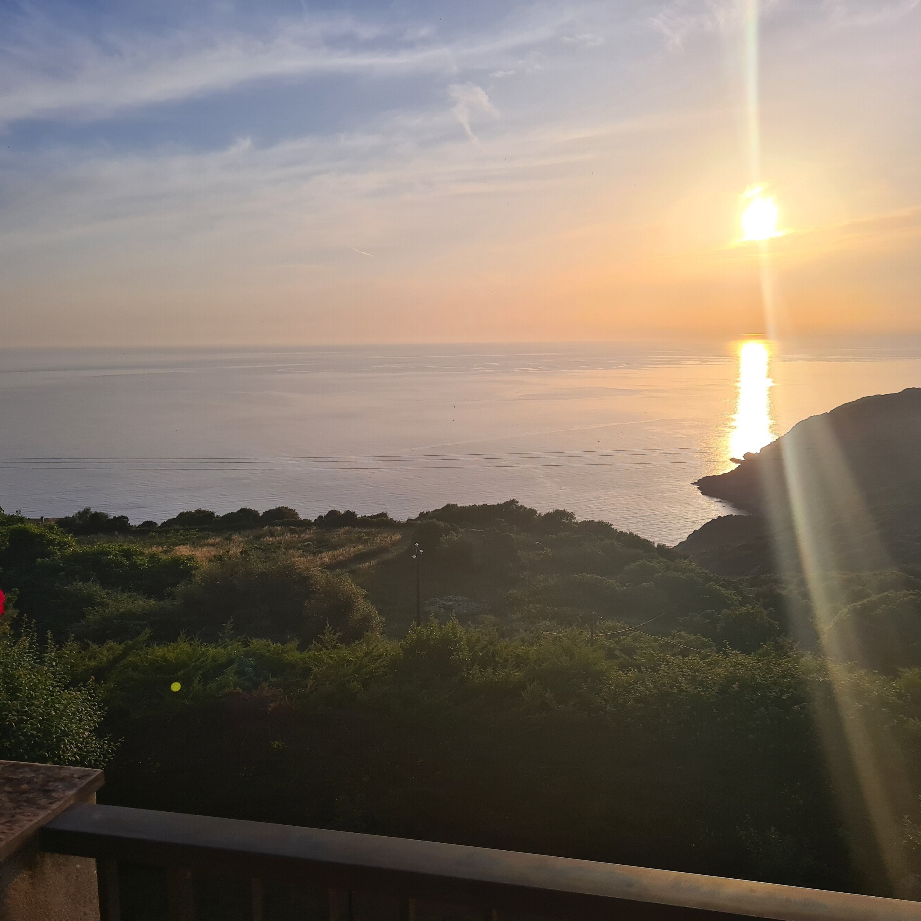 Eine Küstenlandschaft mit Sonnenuntergang über dem Meer, felsigem Gelände und Vegetation.