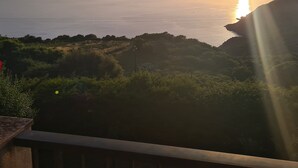 A coastal landscape with a sunset over the sea, rocky terrain, and vegetation.