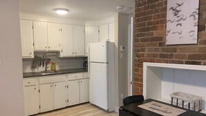Ett modernt kök med vita skåp, en svart bänkskiva och ett vitt kylskåp. Det finns ett matbord med stolar och en vägghylla med en väggmålning som föreställer fåglar.