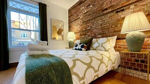 A hotel room with a brick wall, a bed with a patterned bedspread, two bedside lamps, and a window with curtains. - Nice Condo in Limoilou (Quebec City)