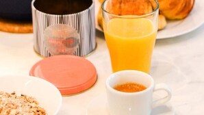 Une table de petit-déjeuner avec un croissant, du jus d’orange, un verre de jus d’orange, un bol de céréales, une tasse de café et un muffin.