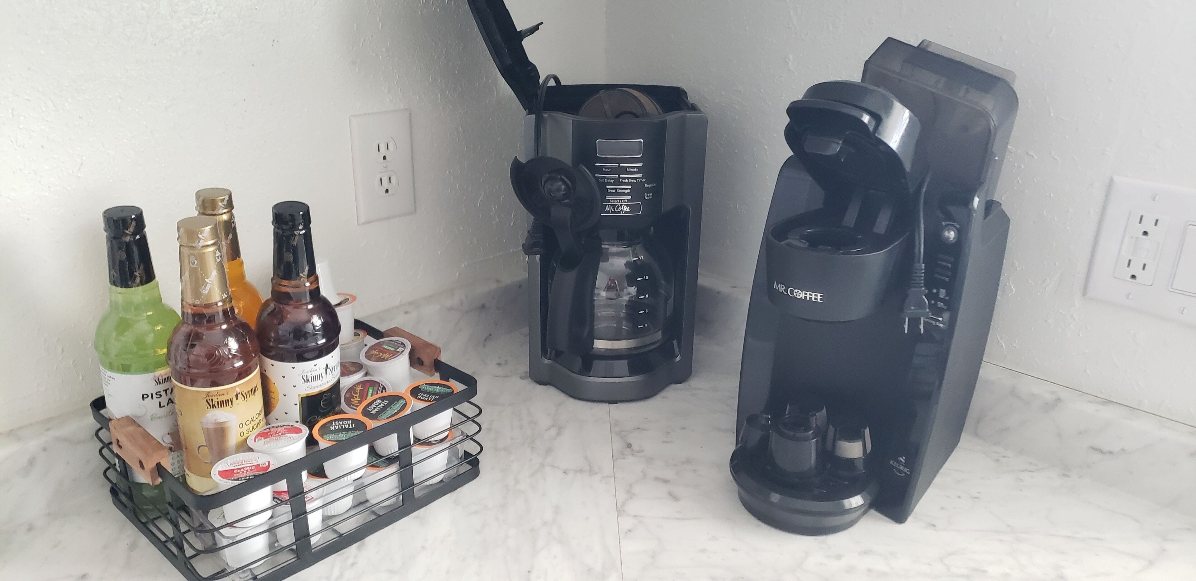 A coffee maker, a basket of coffee pods, and bottles of syrup on a countertop.