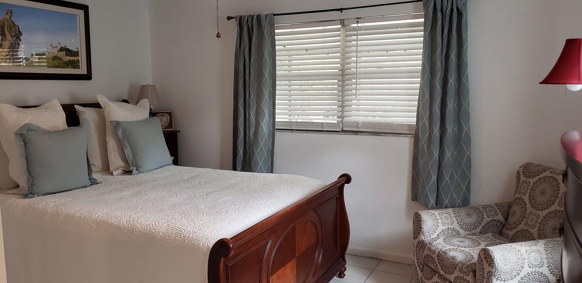 Een slaapkamer met een bed, een stoel, een raam met zonwering en een lamp.