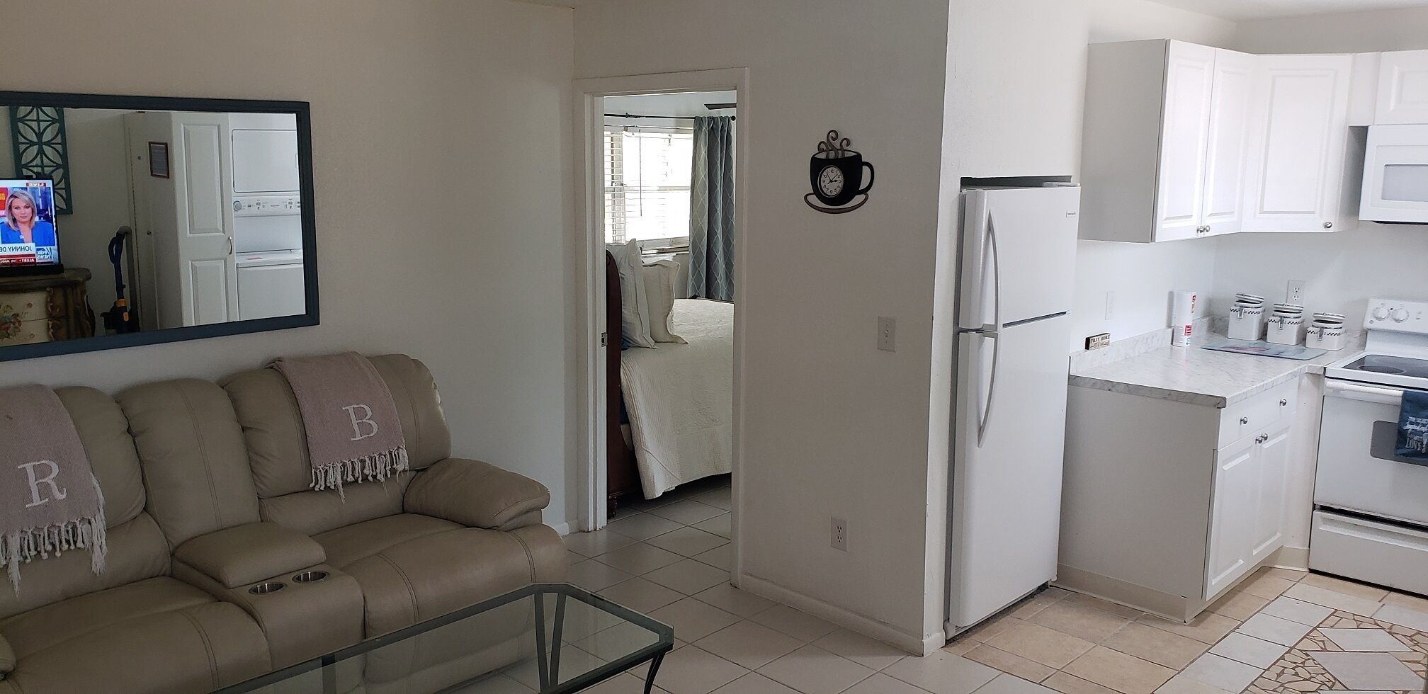 A living room with a beige sofa and a glass coffee table, a kitchen with white appliances, and a hallway leading to a bedroom.