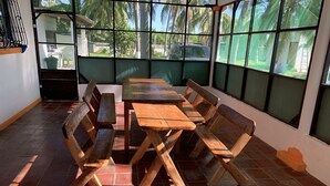 Un área de comedor al aire libre techada con una mesa y bancos de madera, ventanas amplias y vista a palmeras y un coche estacionado.