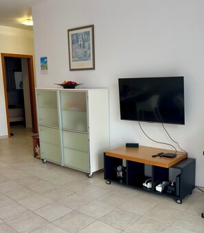 Habitació amb televisió penjada a la paret, un armari blanc amb portes de vidre i una taula petita amb dispositius electrònics.