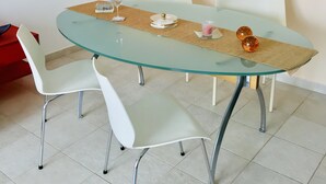 A round glass table with a woven placemat, surrounded by white chairs with metal legs. - Apartment “Golden View” (Porto Santo)