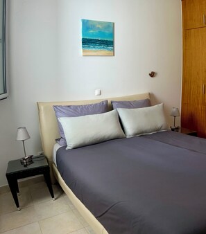 A bedroom with a bed, two bedside tables, a lamp, a painting of a beach scene, and a wardrobe. - Apartment “Golden View” (Porto Santo)