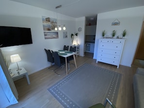 A modern living room with a dining area, a flat-screen TV mounted on the wall, a white cabinet with decorative items, and a grey rug on the floor.