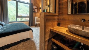 Une chambre aménagée dans une cabane en bois, avec une grande fenêtre, un lit, une coiffeuse en bois dotée d’un lavabo et un miroir.