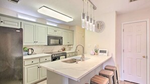 A modern kitchen with white cabinets, stainless steel appliances, a large island with stools, pendant lighting, and a clock on the wall.