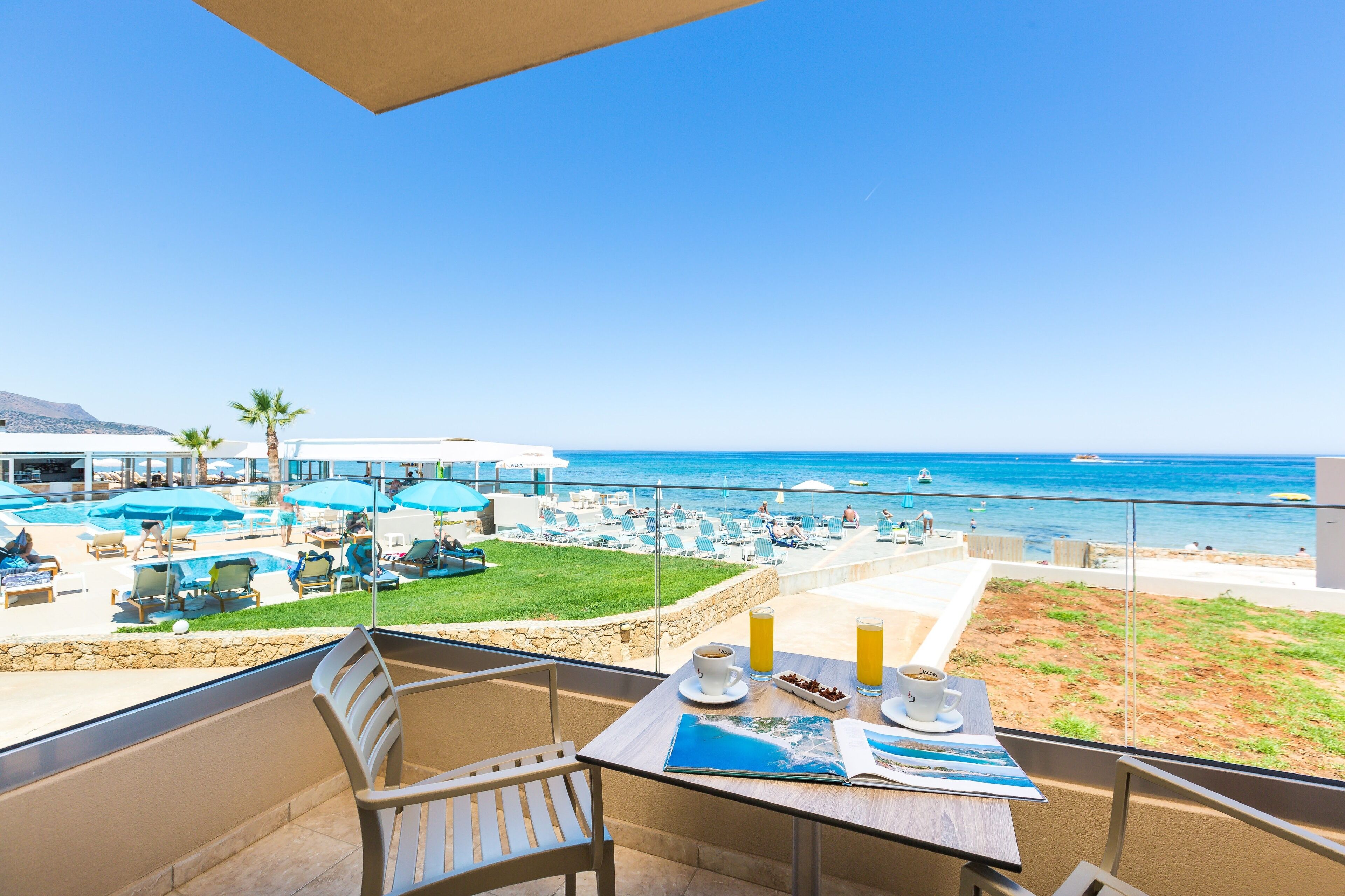 A balcony with a table set for breakfast, overlooking a pool area and beach with umbrellas and lounge chairs.