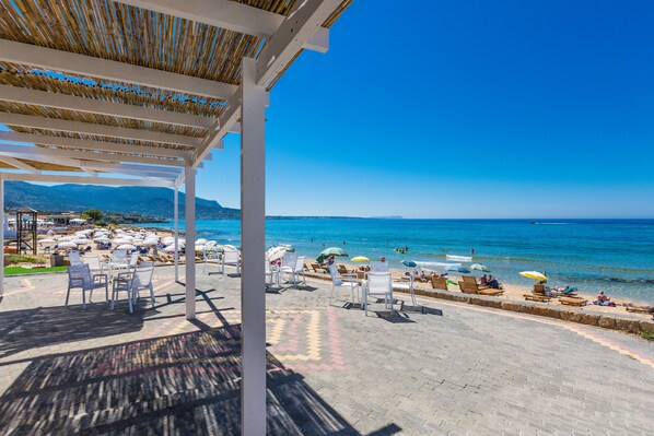 A beachside patio with white chairs and umbrellas, overlooking the sea and mountains.