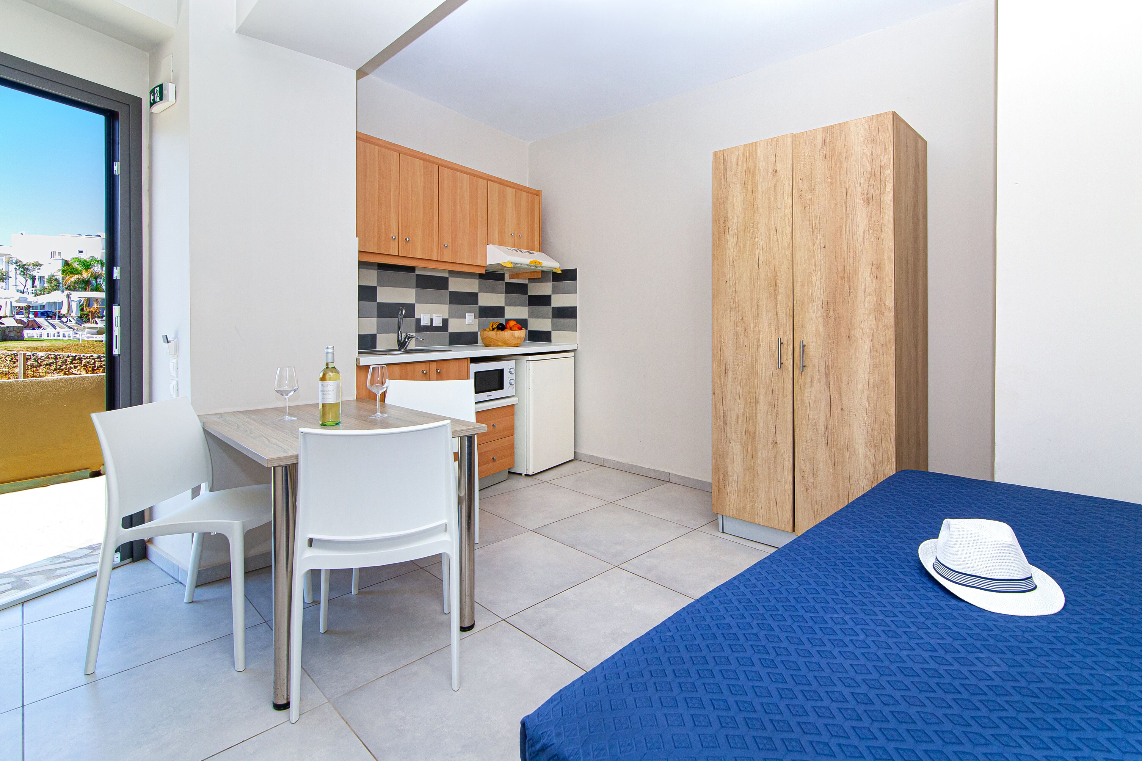 A compact room with a dining table, chairs, a kitchenette, a wardrobe, and a bed with a blue cover and a hat on it.