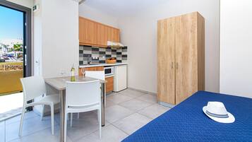 A compact room with a dining table, chairs, a kitchenette, a wardrobe, and a bed with a blue cover and a hat on it.