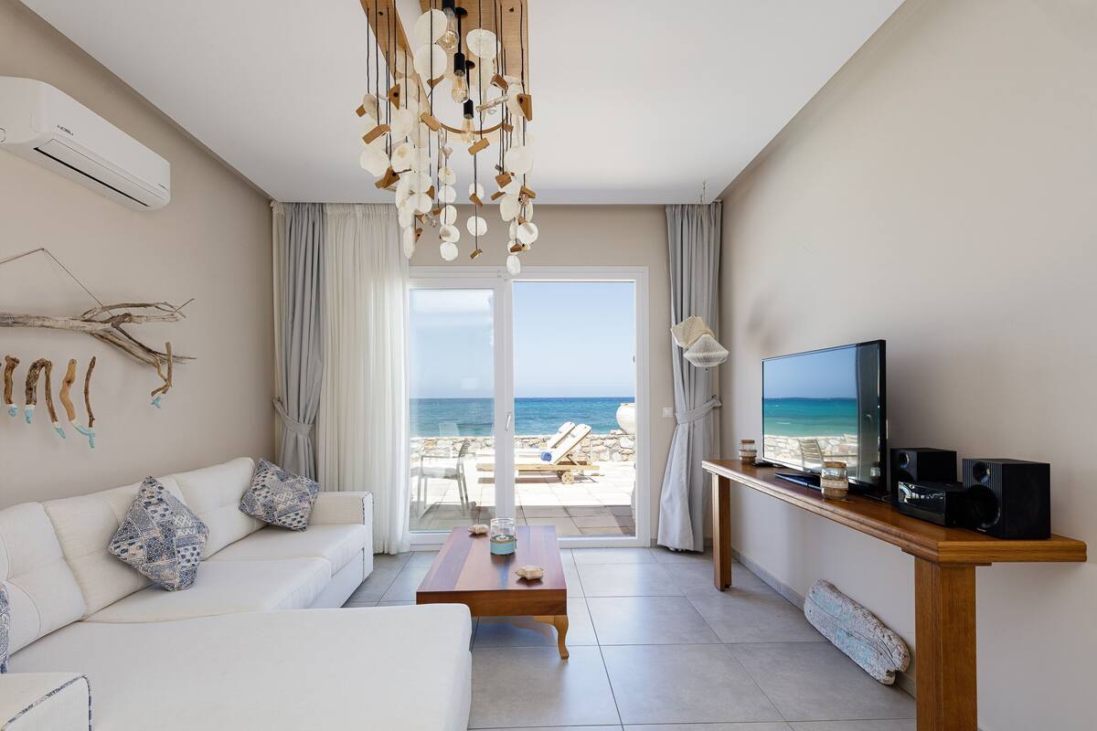 Een moderne woonkamer met een bank, salontafel en televisie. Een groot raam biedt uitzicht op het strand.