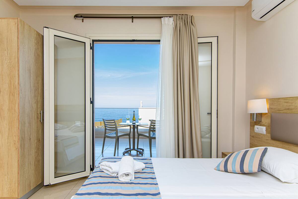 Een hotelkamer met een bed, uitzicht op zee, een balkon met tafel en stoelen, en een schuifdeur.