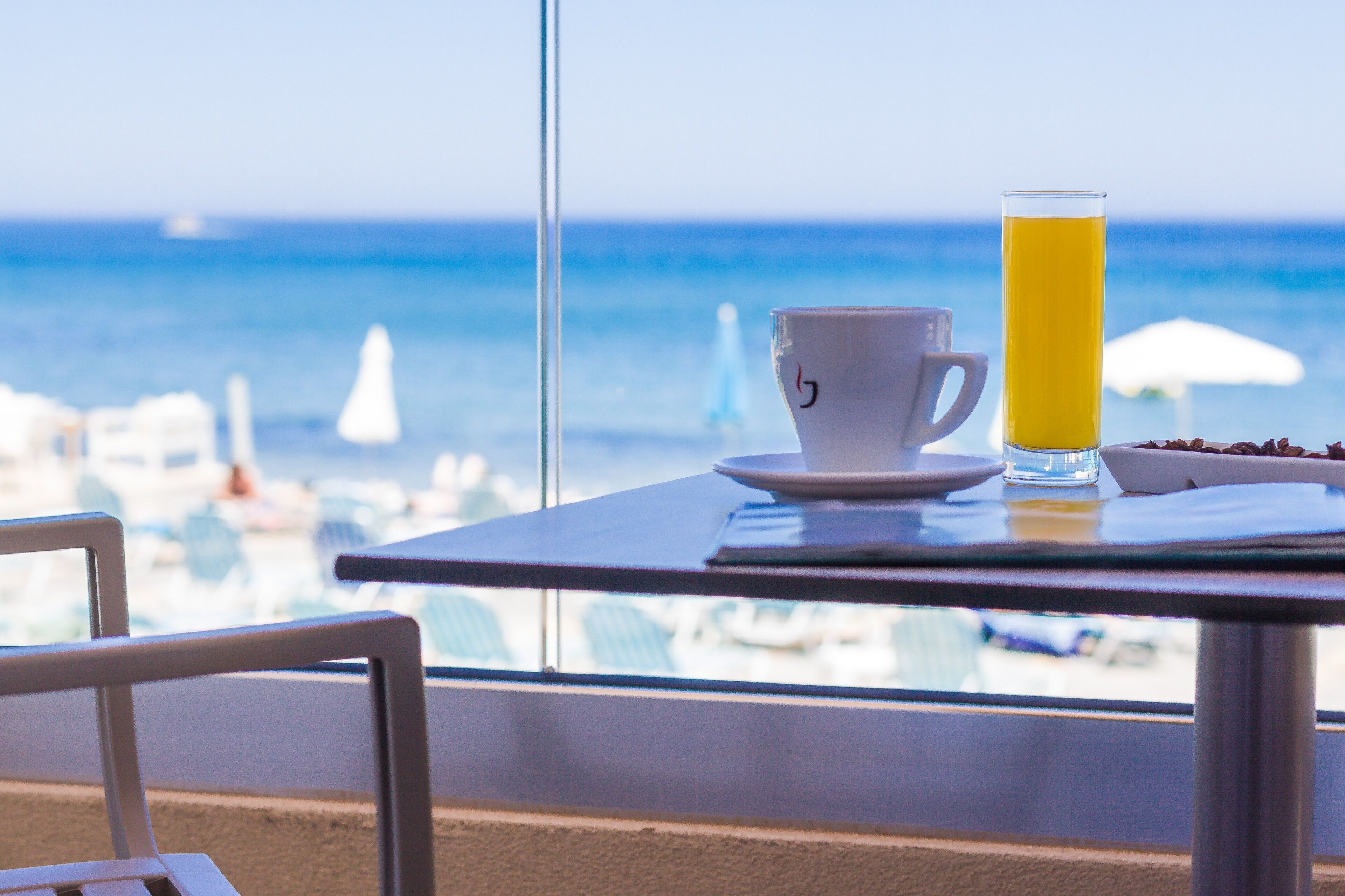 Una mesa con una taza y un vaso alto de jugo de naranja, con vista a una playa con sombrillas y barcos.