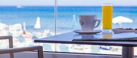 Una mesa con una taza y un vaso alto de jugo de naranja, con vista a una playa con sombrillas y barcos.