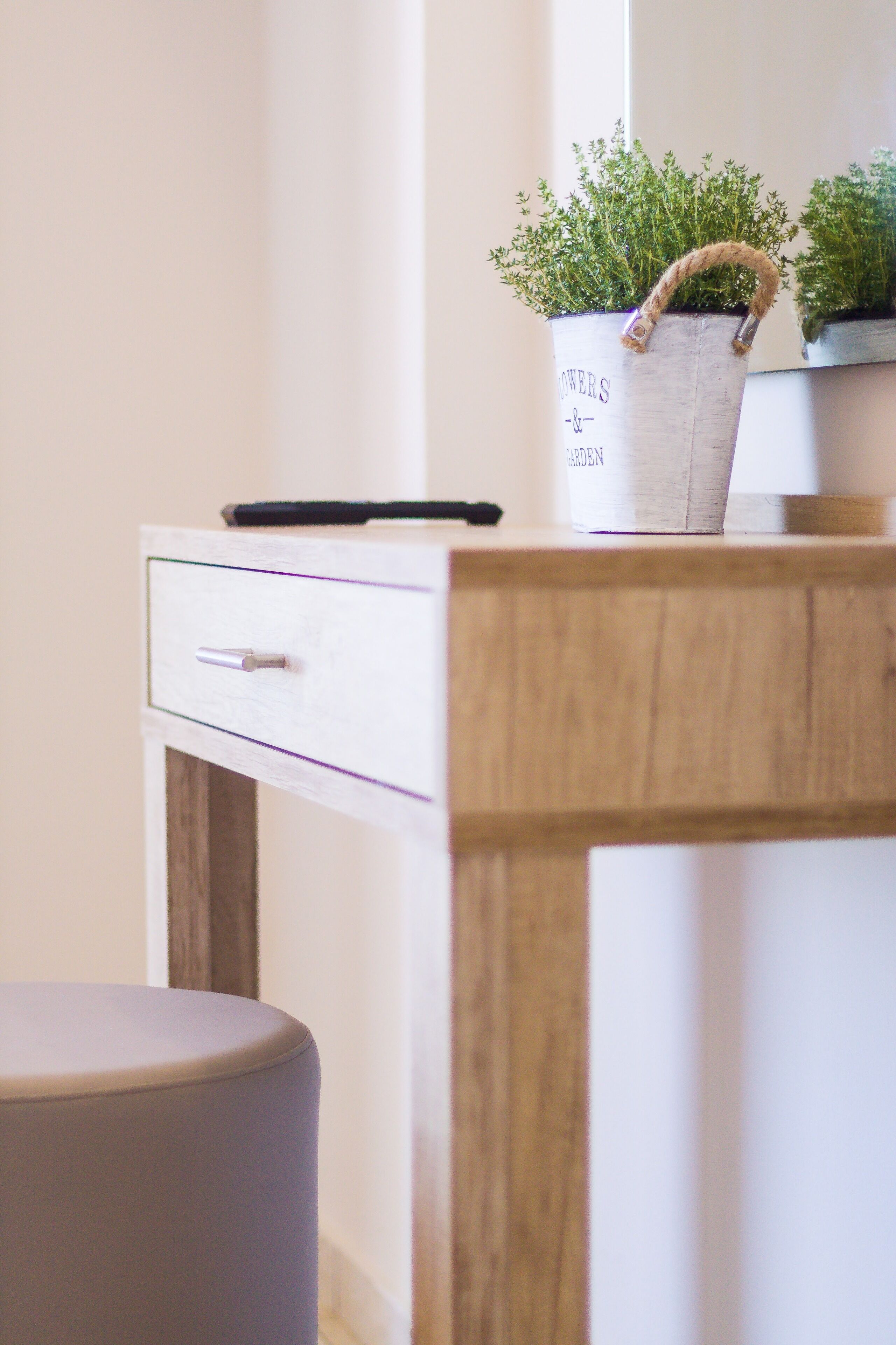 Une table en bois avec un tiroir blanc, un petit tabouret et deux plantes en pot dotées d’une anse en corde.