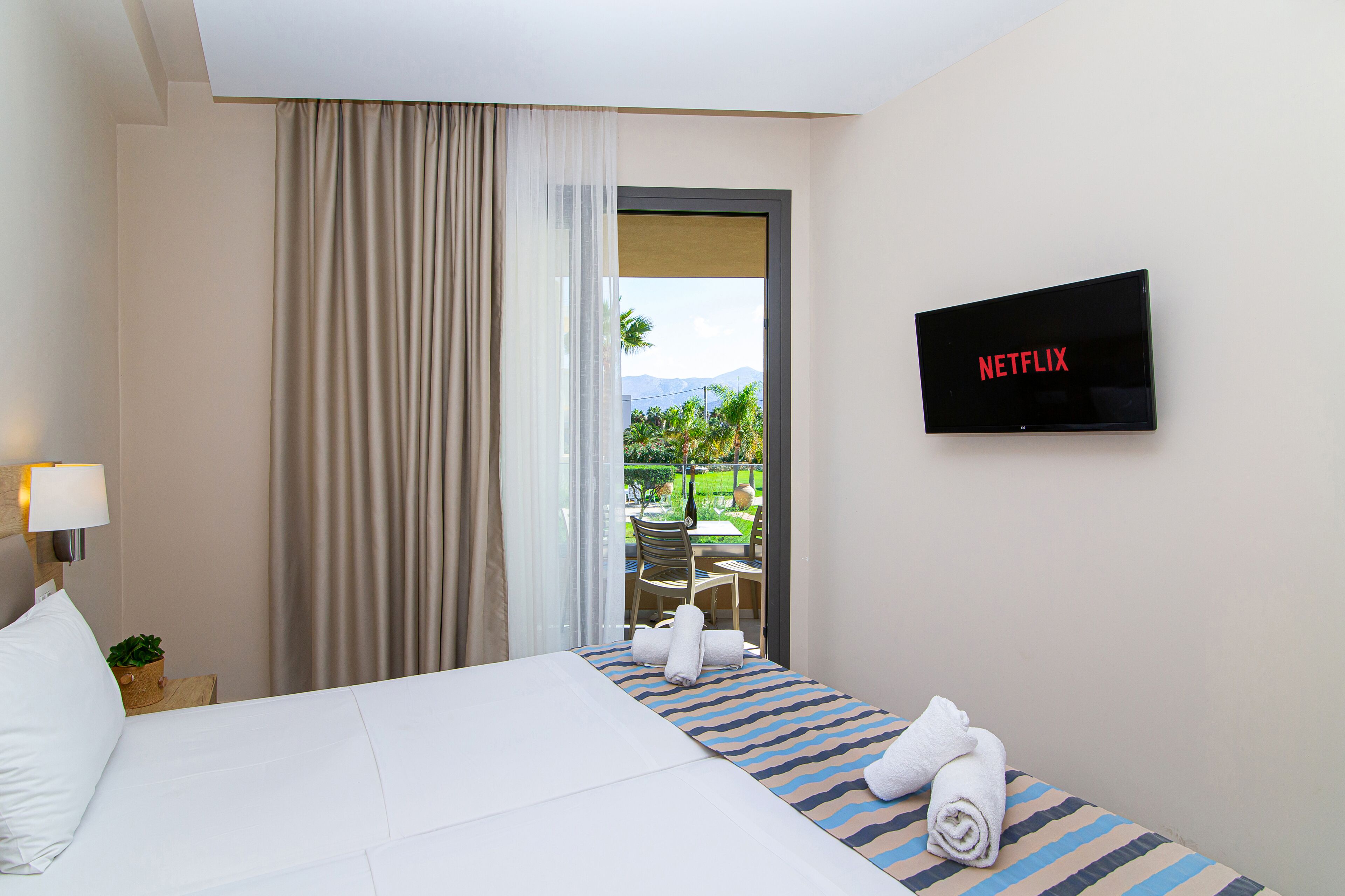 A hotel room with a bed, a TV displaying 'NETFLIX', a window with curtains, and a view of a garden.