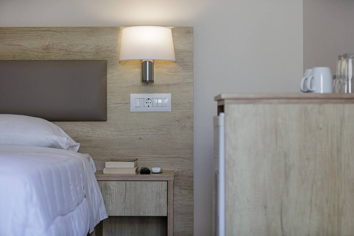 Een opgemaakt bed met witte lakens, een nachtkastje met lamp, een boek en een afstandsbediening.