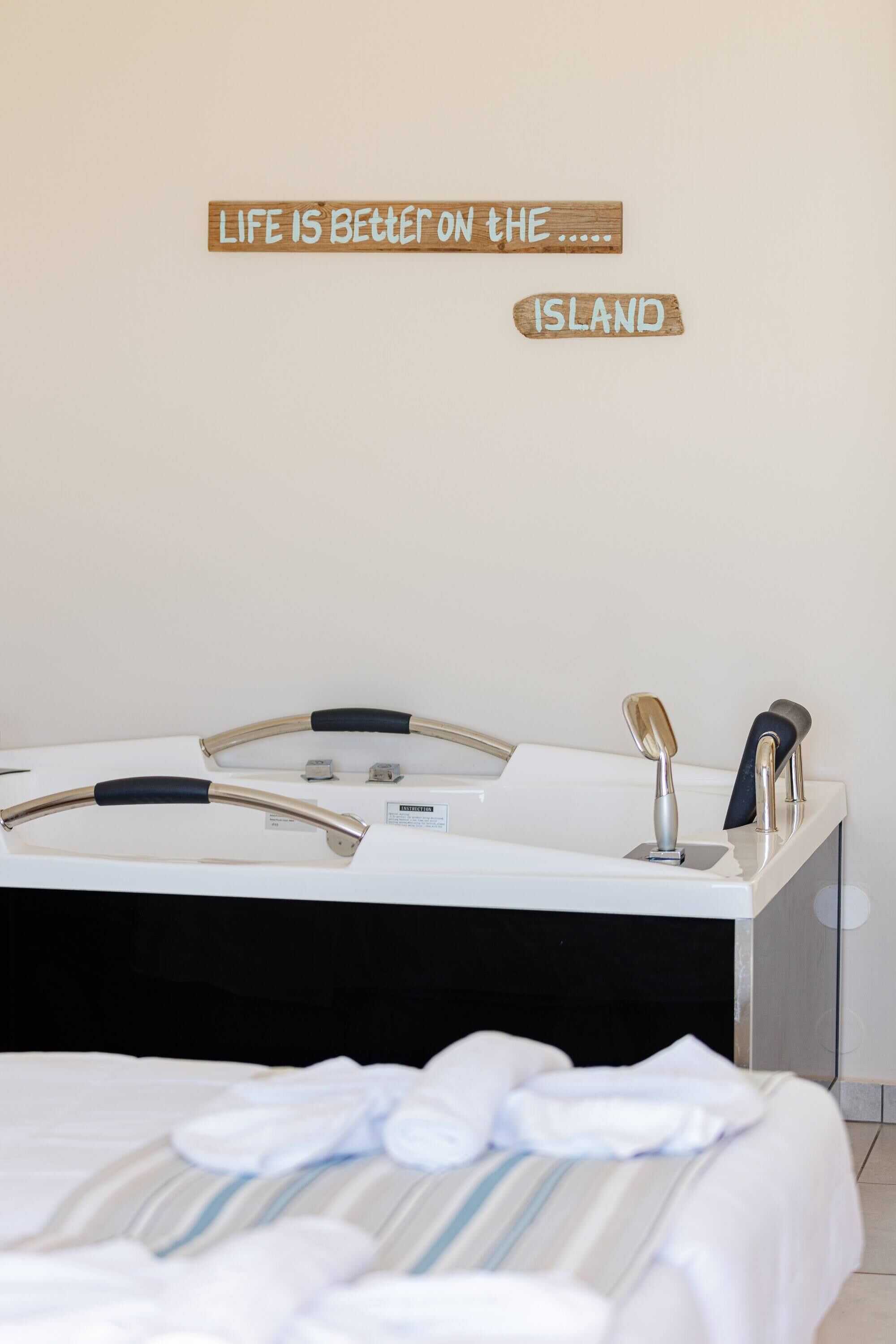A hotel room with a bed, a bathroom with a mirror and towel rack, and a wall with a sign that reads "Life is better on the island."