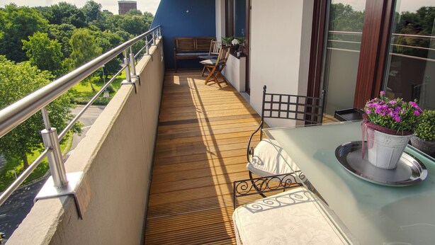 Ein Balkon mit Holzboden, Metallgeländer, einem Tisch mit Topfpflanze und einer Bank.