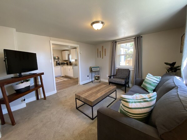 A living room with a television on a wooden stand, a sofa with striped pillows, a coffee table, and a view of a kitchen and dining area through an open doorway.