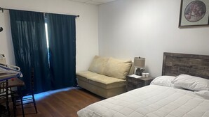 Habitación de hotel con cama, sofá, escritorio con silla, mesita de noche con lámpara y ventana con cortinas.