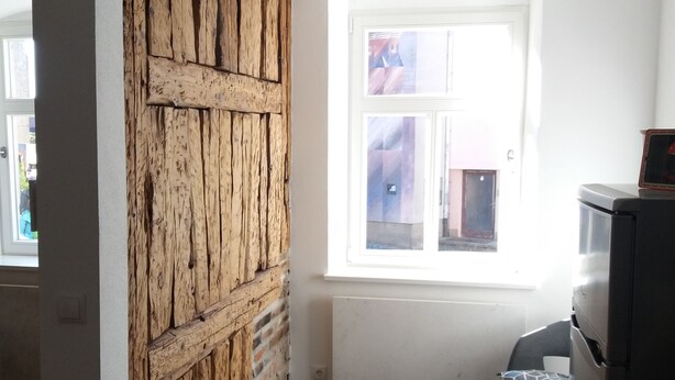 Quarto com parede de madeira rústica, um banquinho preto pequeno, janela com vista e uma área de cozinha com geladeira.