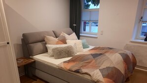 Quarto moderno com sofá cinza, colcha estampada e vista da rua pela janela.