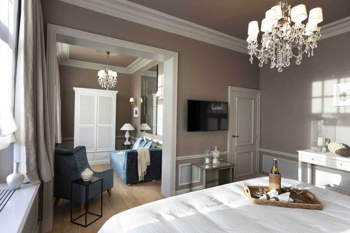A bedroom with a large bed, a TV, a chandelier, and a seating area with a blue sofa and armchairs.