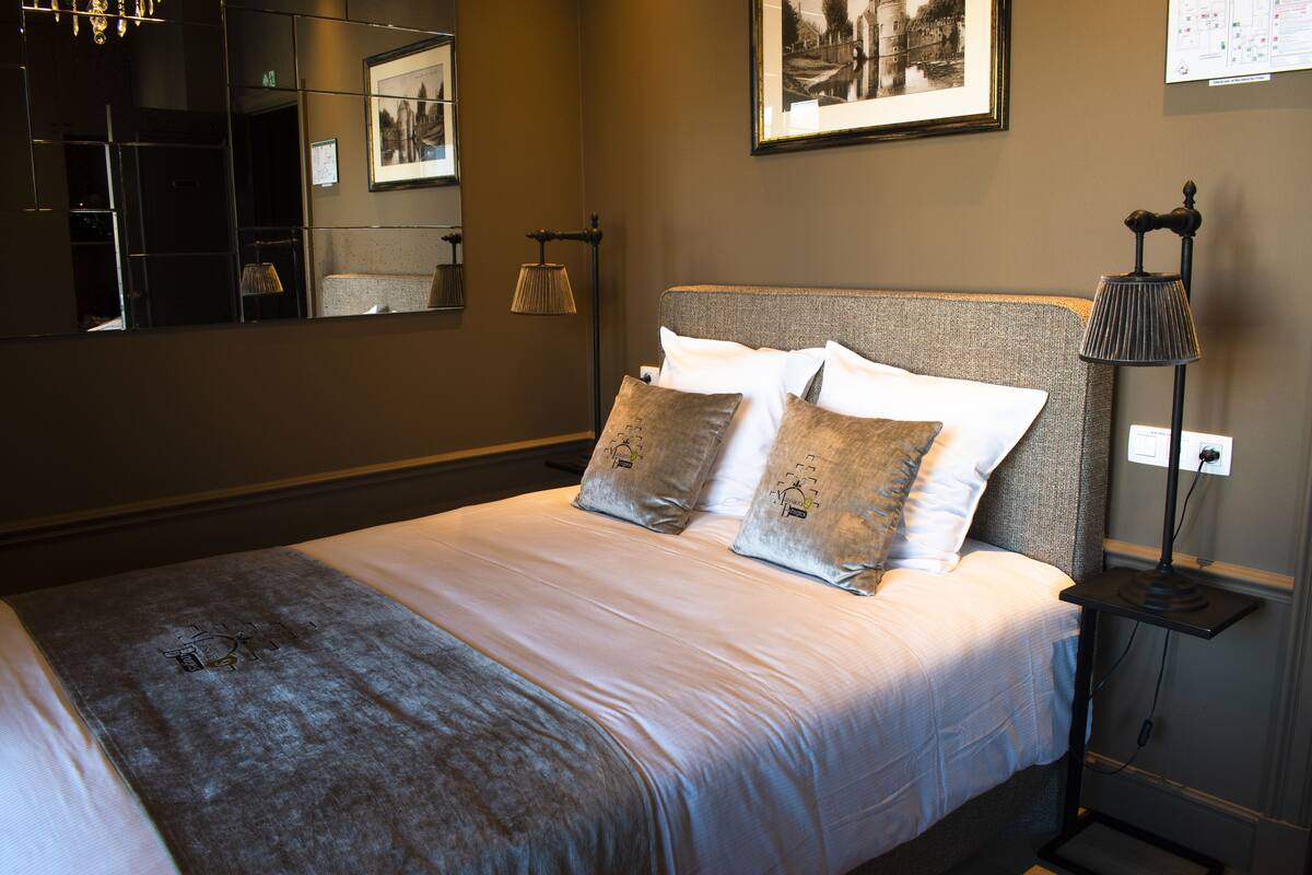 A neatly made bed with a grey headboard, a bedside lamp, and a framed picture on the wall.