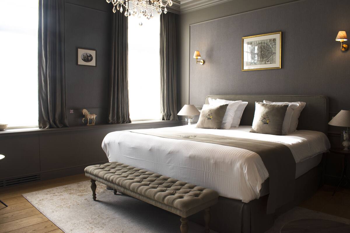 A bedroom with a large bed, a bench, a chandelier, and a window with curtains.
