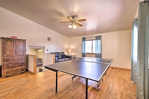 A room with a ping pong table, a ceiling fan, a wooden cabinet, a sofa, and a window with curtains.