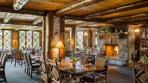 Un coin repas rustique avec des tables et des chaises en bois, une cheminée en pierre et de grandes fenêtres laissant entrer la lumière naturelle.