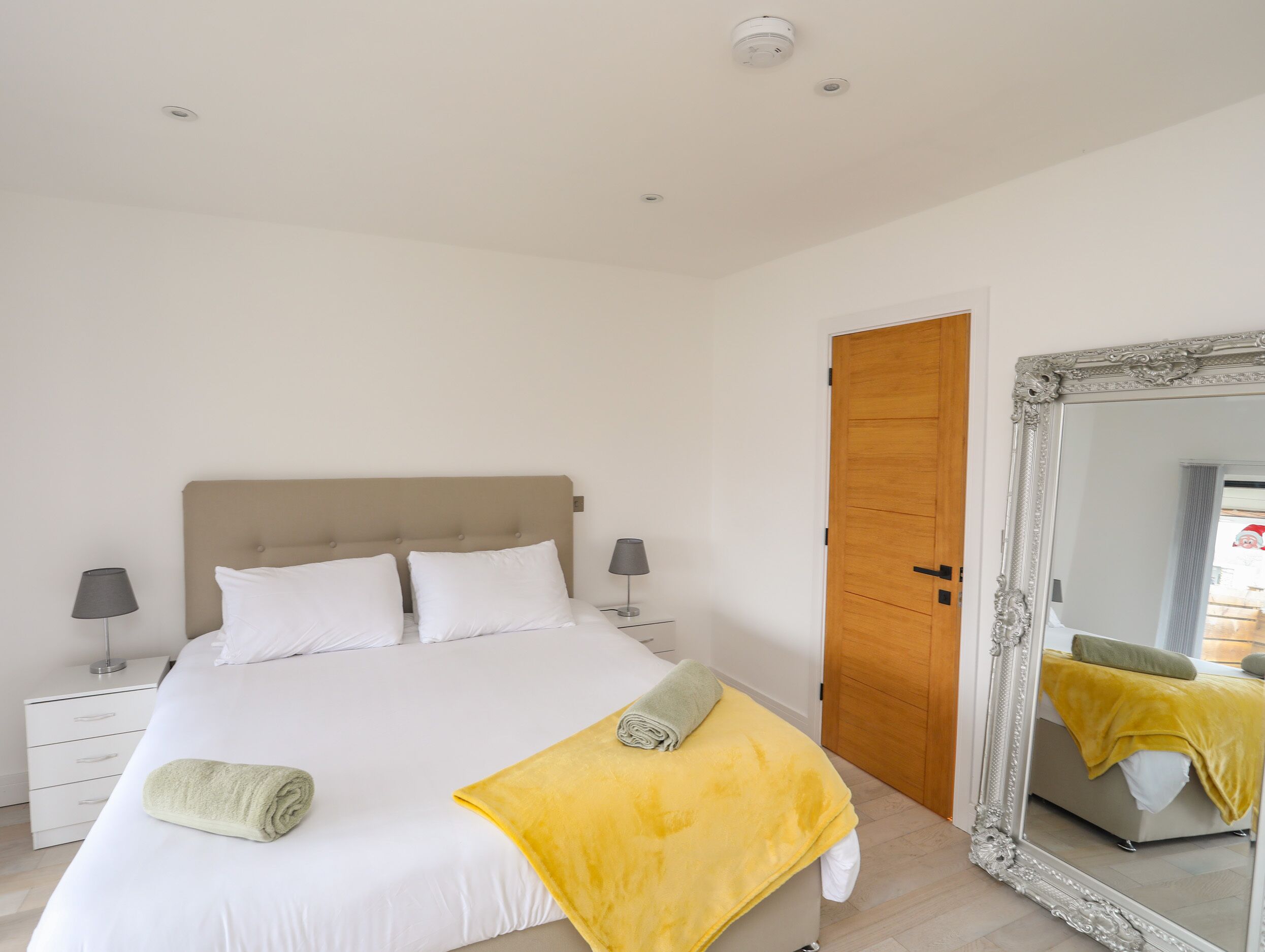 A bedroom with a bed, two bedside tables, a mirror, and a wooden door.