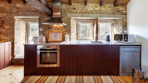Een keuken met een stenen muur, houten balken, een inbouwoven, een vaatwasser en een raam met uitzicht.