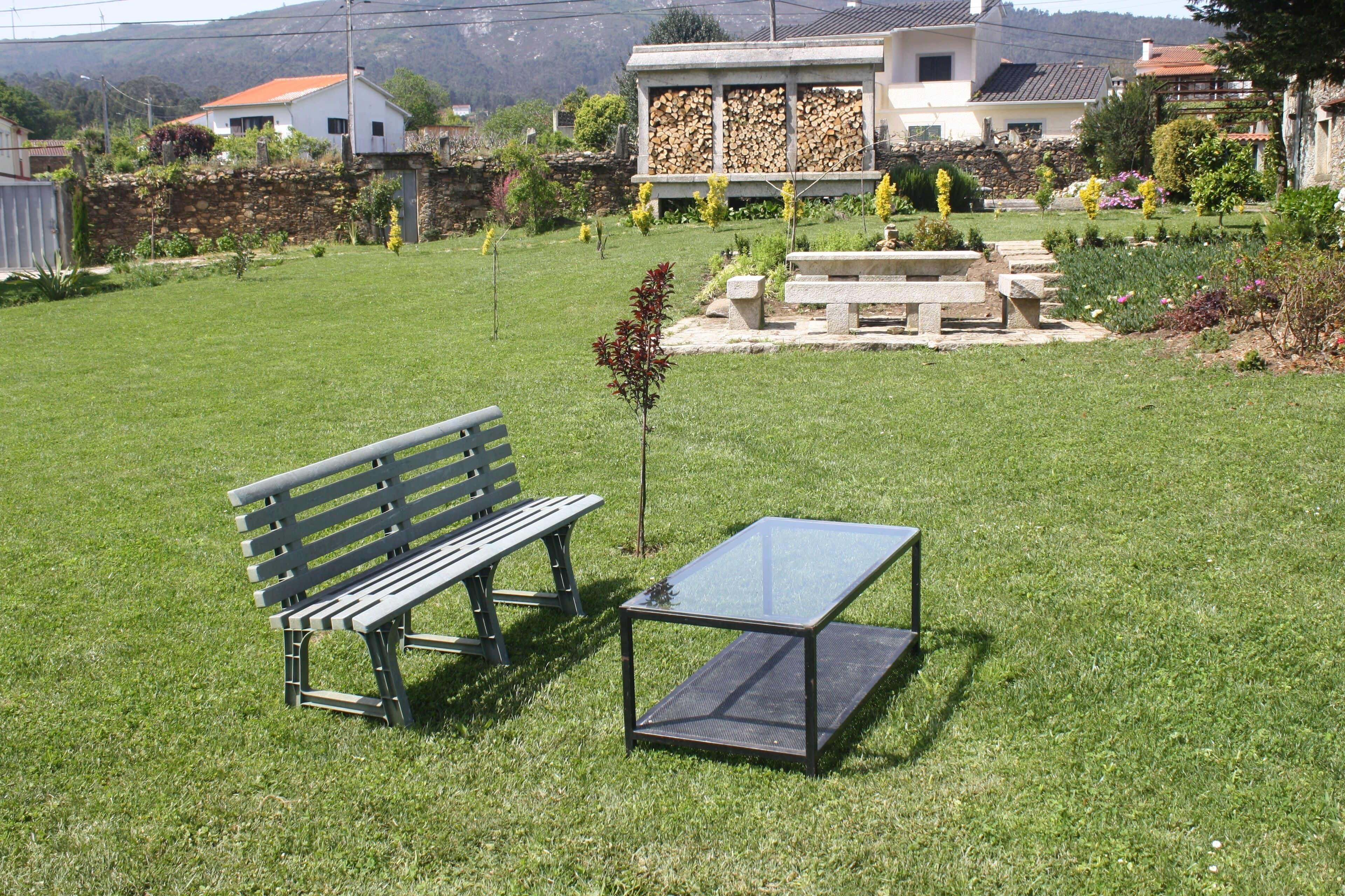 Un espace extérieur verdoyant avec un banc en bois et une table dont le plateau est en verre.