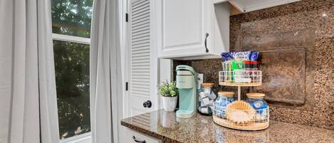 Una cucina con mobili bianchi, un piano di lavoro in granito, una macchina da caffè Keurig, una piccola pianta in vaso e un portaspezie con barattoli.