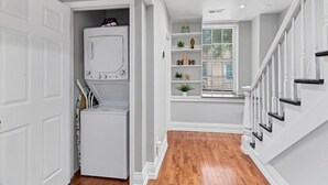 A laundry room with a washer and dryer, a window with blinds, and a staircase with white railings. - The Loft on Main | Steps to Dining & Nightlife (Saint Charles)
