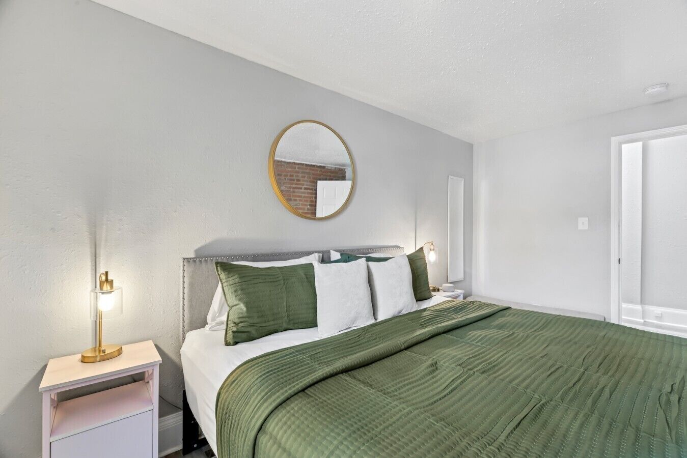A bedroom with a bed, a bedside table, a lamp, a mirror, and a door.
