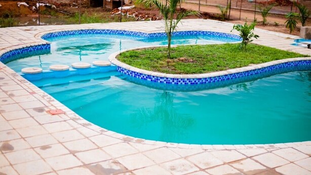 A swimming pool with a tiled edge and a small grassy island in the center. - Self-contained bed sitter overlooking a beautiful scenery in an animal farm. (Lilongwe)