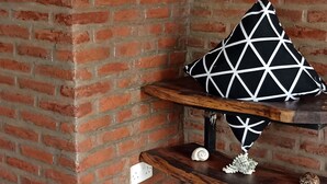 A corner with a brick wall, a wooden shelf with decorative items, and a woven chair with books. - Self-contained bed sitter overlooking a beautiful scenery in an animal farm. (Lilongwe)